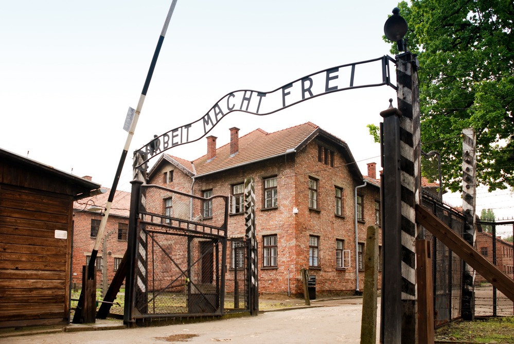 Imagen de la entrada principal del campo de concentración Auschwitz I, con el cartel "Arbeit macht frei" ("El trabajo libera") en la parte superior. El edificio de ladrillo rojo y las instalaciones de alambre de púa son visibles en el fondo. Auschwitz I fue uno de los campos más notorios del régimen nazi, conocido por su historia durante el Holocausto. Hoy en día, es un sitio conmemorativo y museo que recuerda las tragedias ocurridas en este lugar.